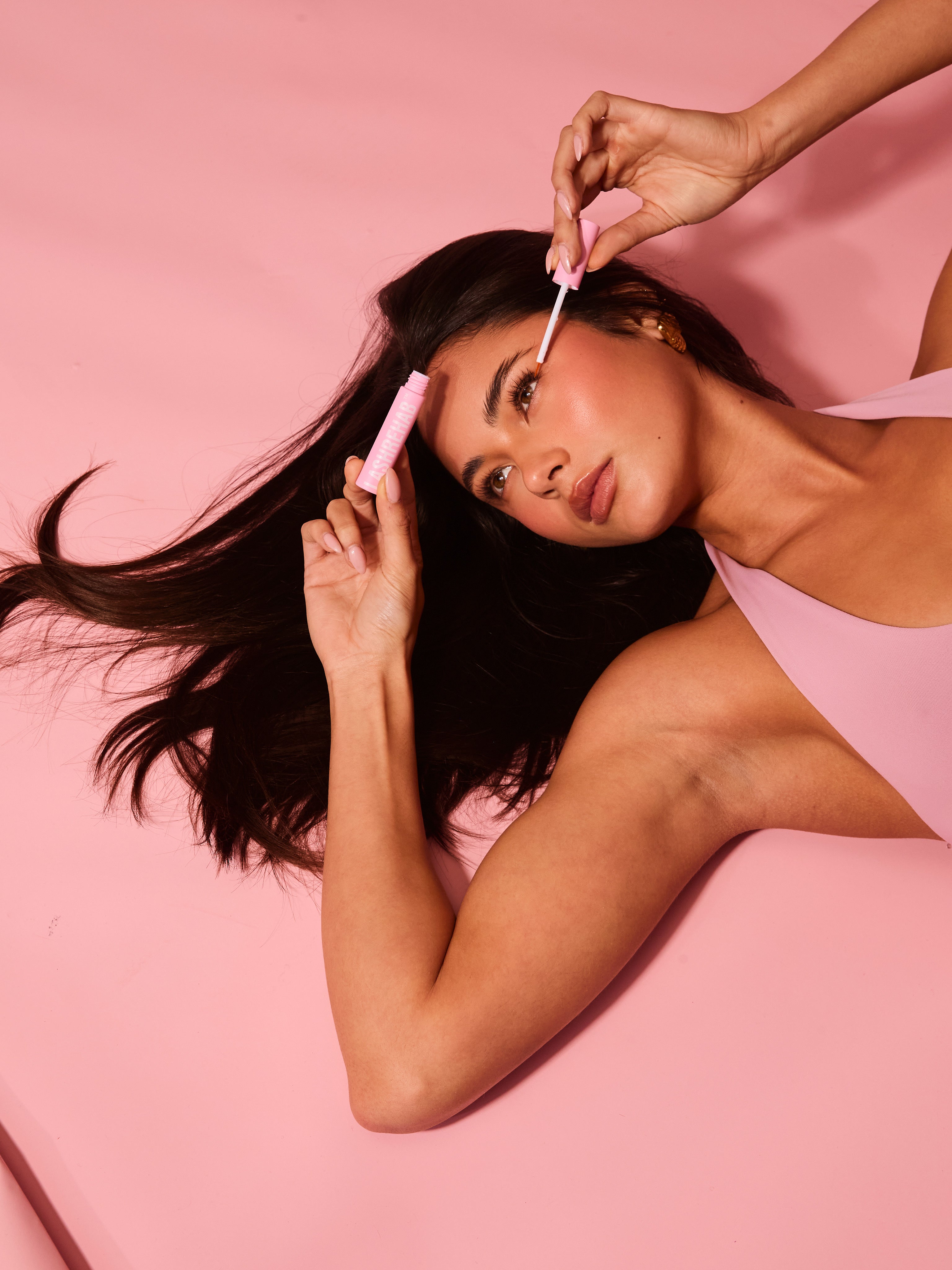 Model applying Lash Rehab lash serum while lying on pink backdrop, demonstrating real use and effortless integration into a daily beauty routine.