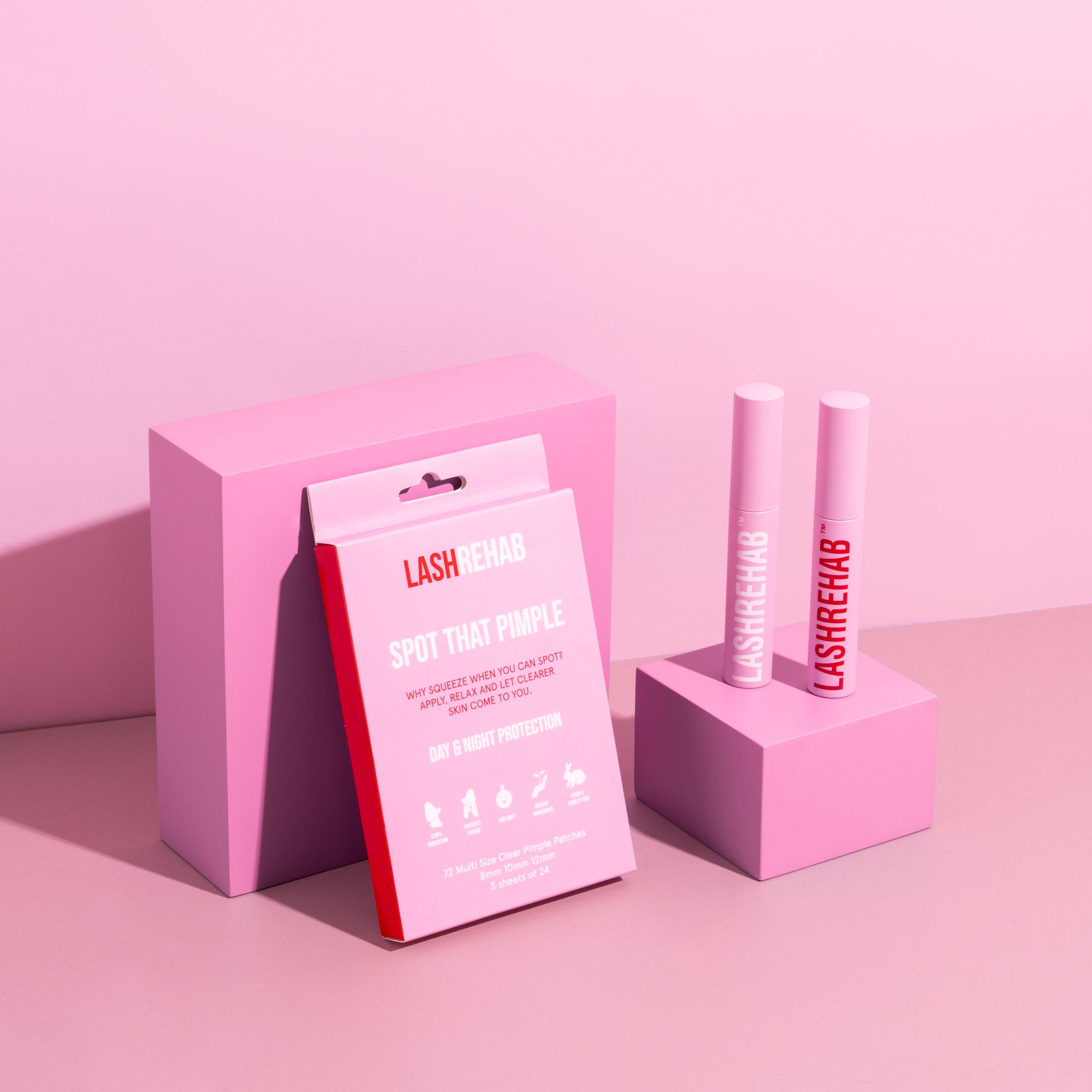 Flat lay of Lash Rehab Spot That Pimple acne patches and two lash applicator tubes styled with pink display blocks