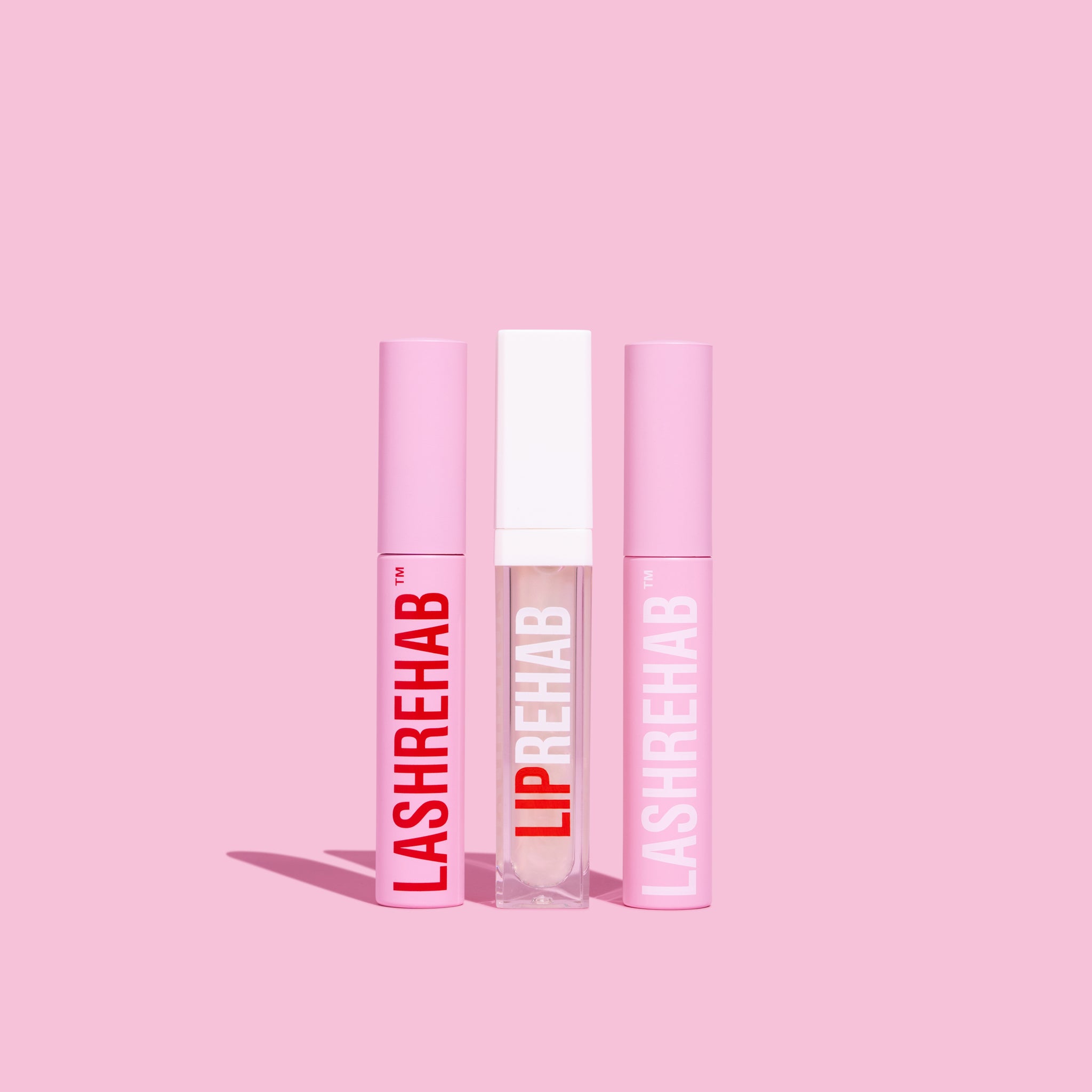 Flat lay of Lash Rehab lash serums and lip serum standing upright on a pink background, showcasing the brand’s signature pink and red minimalist packaging.