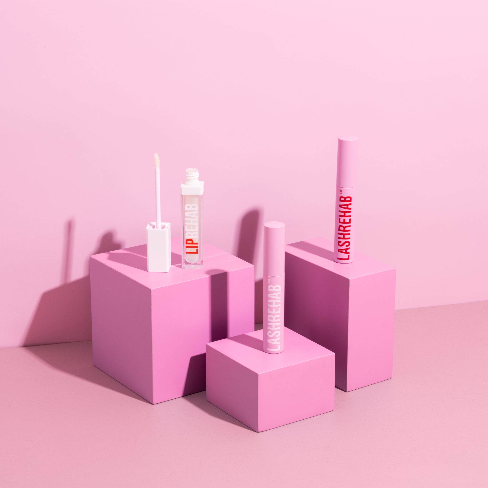 Three Lash Rehab beauty products displayed on pink cube pedestals against a pink backdrop, featuring two pink lash serum tubes and one clear lip serum with wand applicator.